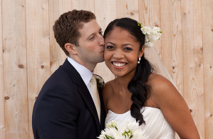 happy white groom kissing black bride holding white folowers